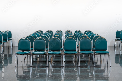 Many empty green leather chairs in a wide hall room, lights on. A floor reflect the objects. Give a feeling of emptiness.