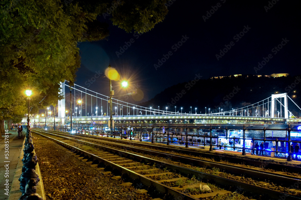 A brightly lit night bridge near a dark mountain. In the foreground are the tram rails in perspective