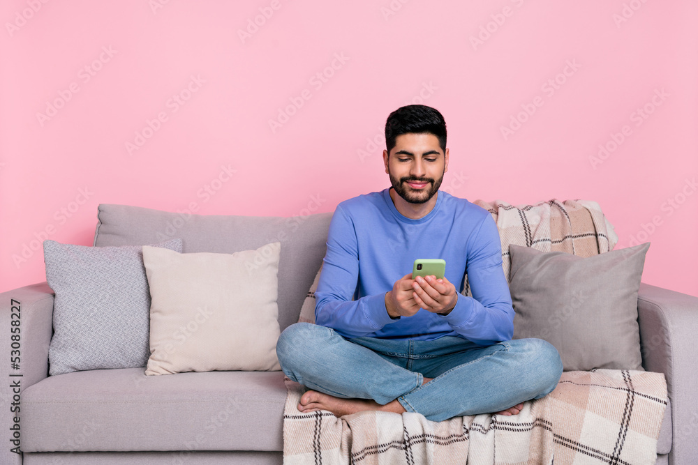 Photo of pretty cute arab guy dressed blue pullover sitting couch typing modern device empty space isolated pink color background Photo of pretty cute arab guy dressed blue pullover sitting couch typing modern device empty space isolated pink color background