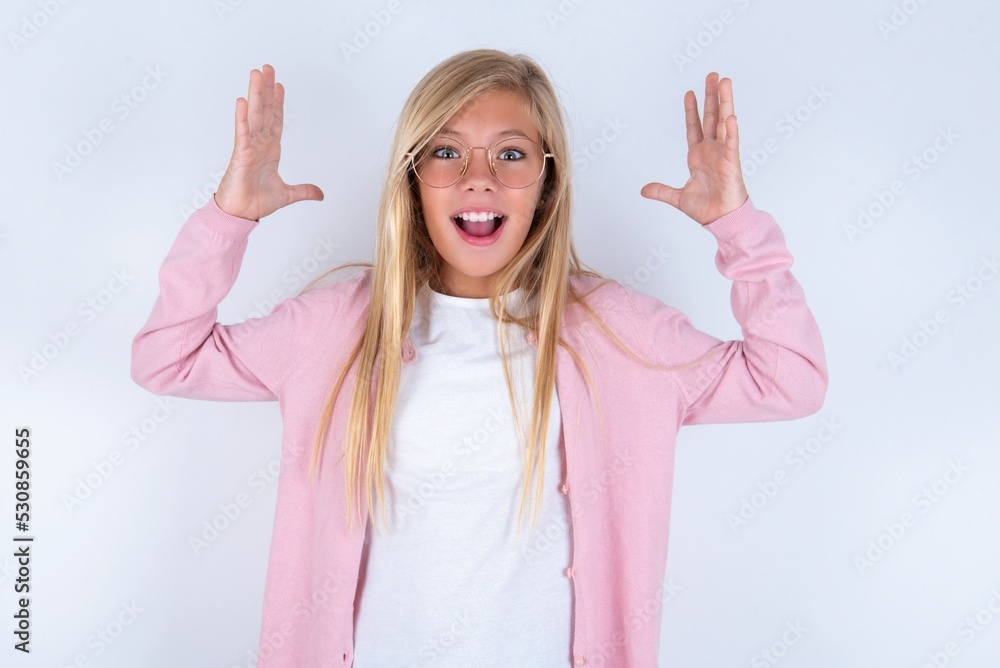 caucasian blonde little girl wearing pink jacket and glasses over white background looks with excitement at camera, keeps hands raised over head, notices something unexpected, reacts on sudden news. caucasian blonde little girl wearing pink jacket and glasses over white background looks with excitement at camera, keeps hands raised over head, notices something unexpected, reacts on sudden news.