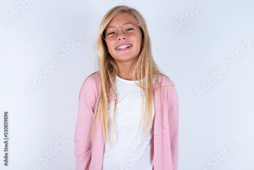 caucasian blonde little girl wearing pink jacket and glasses over white background with broad smile  shows white teeth  feeling confident rejoices having day off.