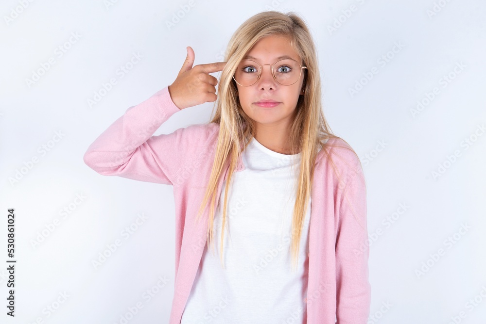 Unhappy caucasian blonde little girl wearing pink jacket and glasses over white background curves lips and makes suicide gesture, shoots in temple with hand, tries kill himself. Unhappy caucasian blonde little girl wearing pink jacket and glasses over white background curves lips and makes suicide gesture, shoots in temple with hand, tries kill himself.