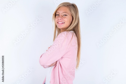 Image of cheerful caucasian blonde little girl wearing pink jacket and glasses over white background with arms crossed. Looking and smiling at the camera. Confidence concept.