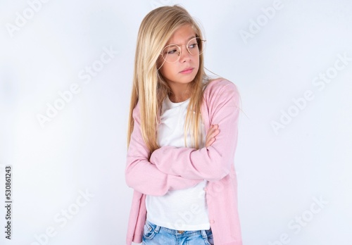 Displeased caucasian blonde little girl wearing pink jacket and glasses over white background with bad attitude, arms crossed looking sideways. Negative human emotion facial expression feelings.