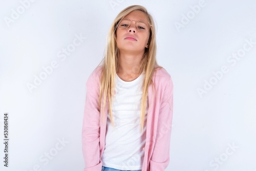 Gloomy  bored caucasian blonde little girl wearing pink jacket and glasses over white background frowns face looking up  being upset with so much talking hands down  feels tired and wants to leave.