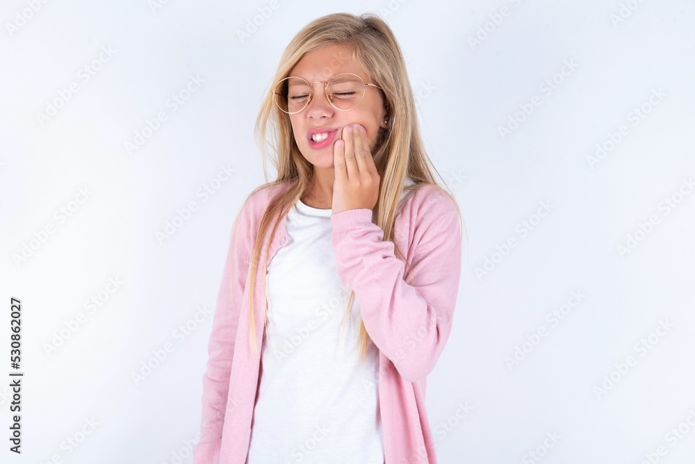 Tooth ache concept. caucasian blonde little girl wearing pink jacket and glasses feeling pain, holding his cheek with hand, suffering from bad toothache, looking at camera with painful expression Tooth ache concept. caucasian blonde little girl wearing pink jacket and glasses feeling pain, holding his cheek with hand, suffering from bad toothache, looking at camera with painful expression