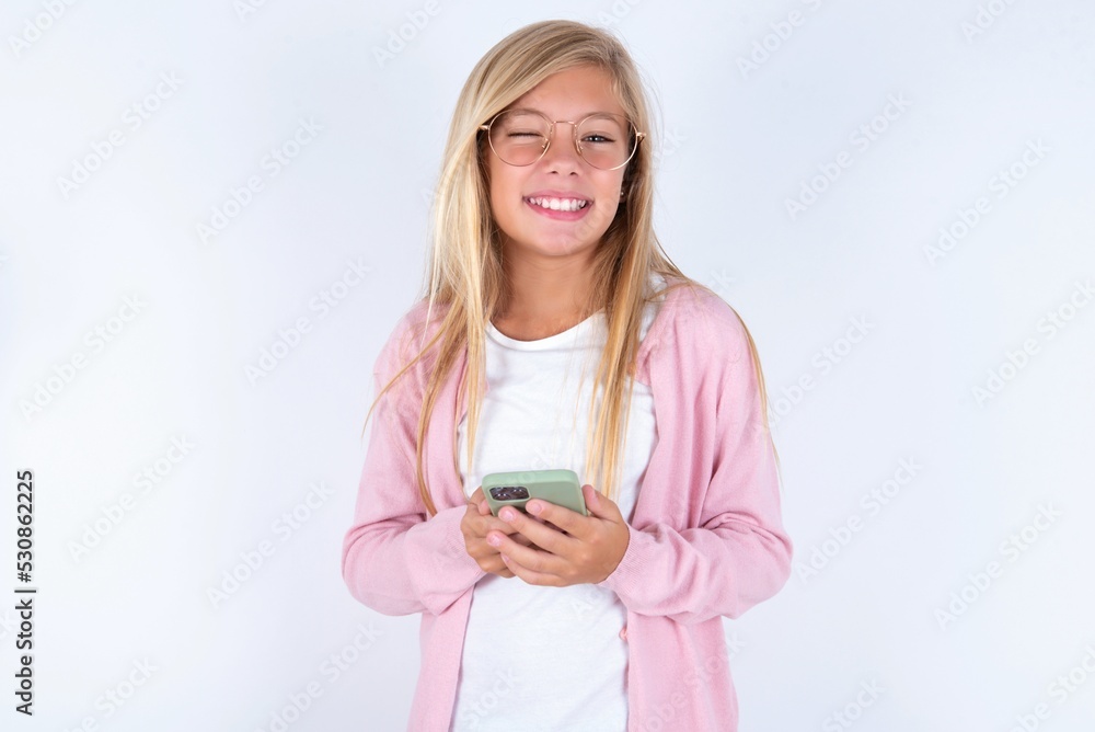 Pleased caucasian blonde little girl wearing pink jacket and glasses over white background using self phone and looking and winking at the camera. Flirt and coquettish concept. Pleased caucasian blonde little girl wearing pink jacket and glasses over white background using self phone and looking and winking at the camera. Flirt and coquettish concept.