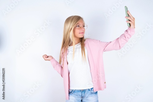 Portrait of a caucasian blonde little girl wearing pink jacket and glasses over white background taking a selfie to send it to friends and followers or post it on his social media.