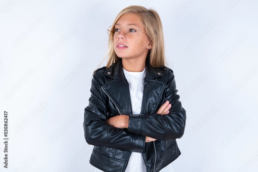Charming thoughtful beautiful caucasian blonde little girl wearing biker jacket and glasses over white wall stands with arms folded concentrated somewhere with pensive expression thinks what to do Charming thoughtful beautiful caucasian blonde little girl wearing biker jacket and glasses over white wall stands with arms folded concentrated somewhere with pensive expression thinks what to do
