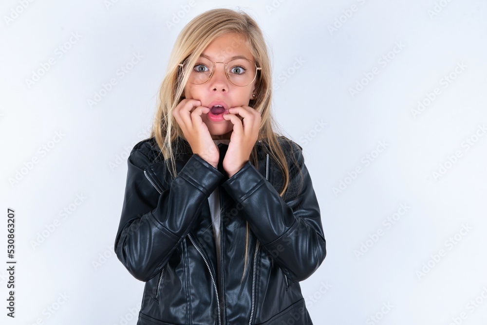 Speechless beautiful caucasian blonde little girl wearing biker jacket and glasses over white background keeps hands near opened mouth reacts to shocking news stares wondered at camera Speechless beautiful caucasian blonde little girl wearing biker jacket and glasses over white background keeps hands near opened mouth reacts to shocking news stares wondered at camera