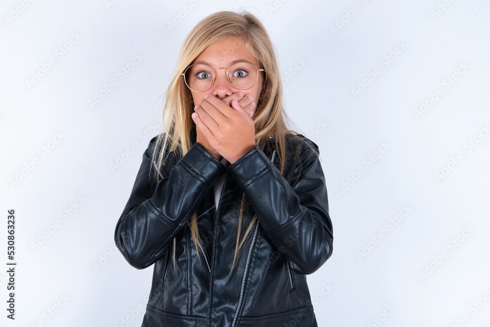 Stunned beautiful caucasian blonde little girl wearing biker jacket and glasses over white background covers mouth with both hands being afraid from something or after hearing stunning gossips. Stunned beautiful caucasian blonde little girl wearing biker jacket and glasses over white background covers mouth with both hands being afraid from something or after hearing stunning gossips.