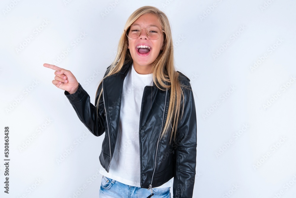 beautiful caucasian blonde little girl wearing biker jacket and glasses over wh laughs happily points away on blank space demonstrates shopping discount offer, excited by good news or unexpected sale. beautiful caucasian blonde little girl wearing biker jacket and glasses over wh laughs happily points away on blank space demonstrates shopping discount offer, excited by good news or unexpected sale.