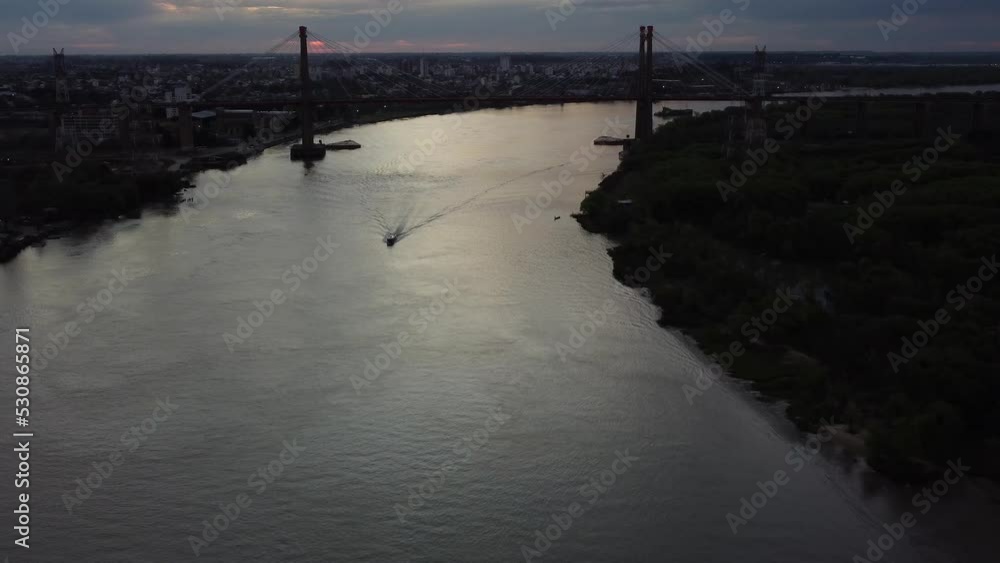 A small motorized boat speeding along a big river underneath a spanned metal brings towards the camera a sunset Aerial video A small motorized boat speeding along a big river underneath a spanned metal brings towards the camera a sunset Aerial video