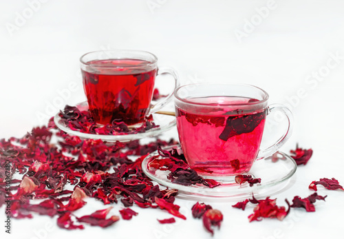 Two cups of Red hot hibiscus tea in a glass mug on a white table among dry Sudanese rose flowers.