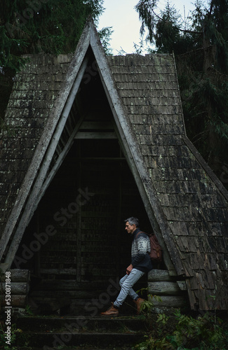 Men traveler with backpack resting inside old wooden dark cabin among woodland. Dark vintage style image