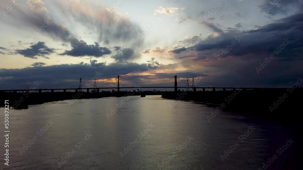 Camera following along a big river towards a spanned metal bridge at sunset with ominous clouds on the horizon Camera following along a big river towards a spanned metal bridge at sunset with ominous clouds on the horizon