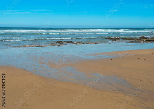 View of empty Praia do Queimado beach with ocean waves and sharp rocks and wet golden sand at wild Rota Vicentina coast near Porto Covo, Portugal.