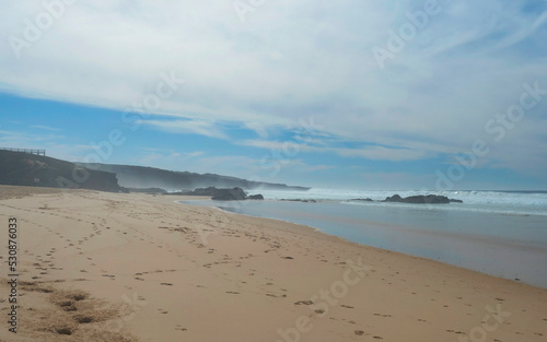View of Praia dos Aivados sand beach with ocean waves and sharp rock and distant figures at wild Rota Vicentina coast near Porto Covo, Portugal.
