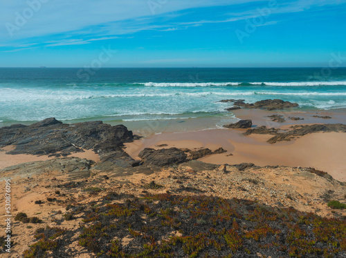 View of empty small sand beach with ocean waves and sharp rocks, stones and green vegetation at wild Rota Vicentina coast near Porto Covo, Portugal.