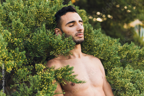 Muscular naked ethnic guy lying on green bush with closed eyes