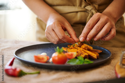 Close-up of serving dish. Selective focus on the hands of a chef putting a rosemary leaf on the top of roasted wedges of organic batata, while preparing healthy delicious vegan meal for dinner