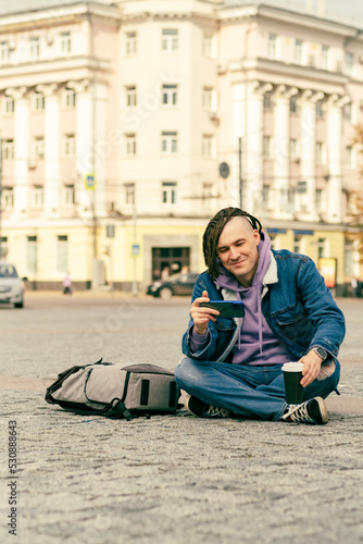 Hipster man with dreadlocks in a denim warm jacket sitting outside on street together with cell phone, and looks at the video content on the smartphone and drinks coffee. Outdoors city background.