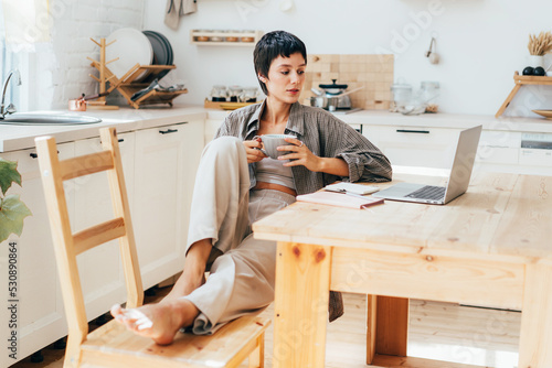 Tablou pe pânză Young female freelancer in the kitchen taking a coffee break while working remotely on a laptop