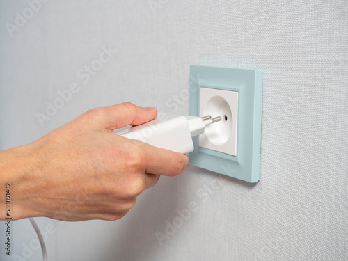 Close-up of a woman's hand holding a white plug and about to plug it into a blue outlet on the wall in the house. The concept of the need for electricity in the house. Mobile devices, Electrification