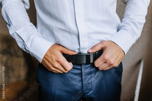 A man, a groom, a businessman getting ready in the morning, fastening a leather black belt on his trousers with his hands. Photography, business.
