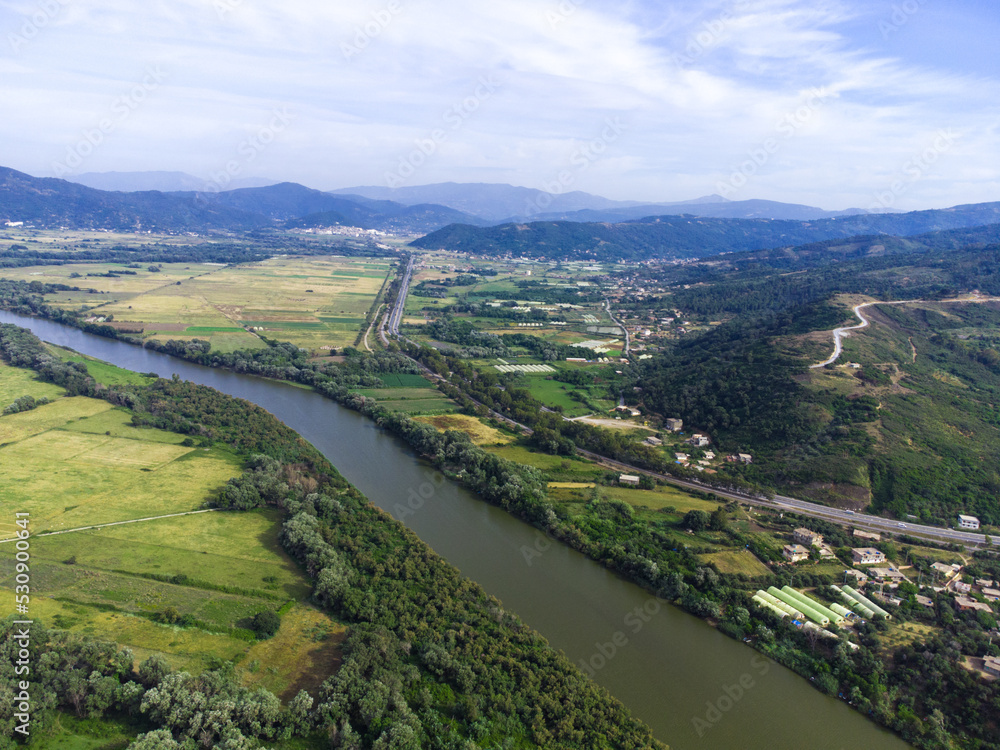 Aerial View of green and yellow agricultural fields, plains with a river in the middle, mountains and cloudy sky in the background, Aerial view of bright river flowing through green meadows, Algeria