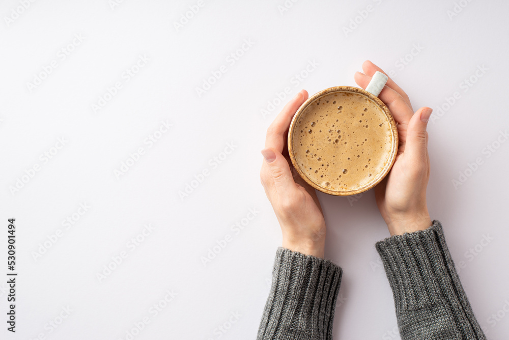 Fototapeta premium Autumn aesthetic concept. First person top view photo of woman's hands in grey sweater touching cup of frothy coffee on isolated white background with empty space