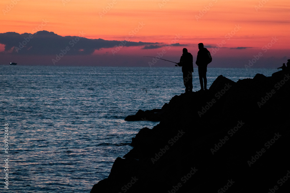 Silhouettes of two fishermen fishing on rocks at sunset. Civitavecchia (Rome) Italy