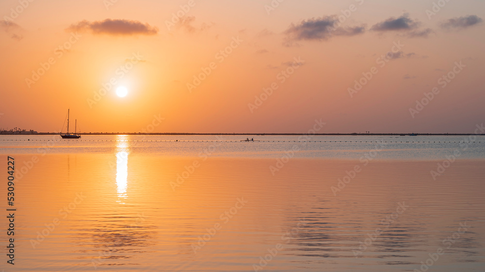 A yellow disk of sun rising into a orange sky, over a calm Mediterranean sea, reflected in the water next to a single sail boat