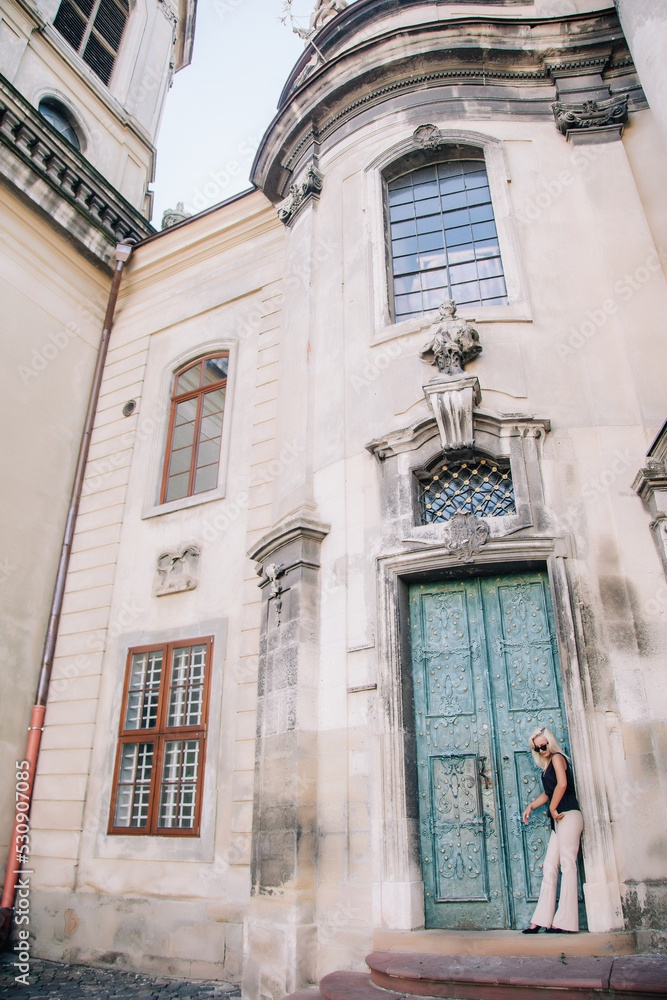Fototapeta premium A young, blonde girl in a black blouse and sunglasses poses against the backdrop of an old courtyard in Lviv. Ukraine.