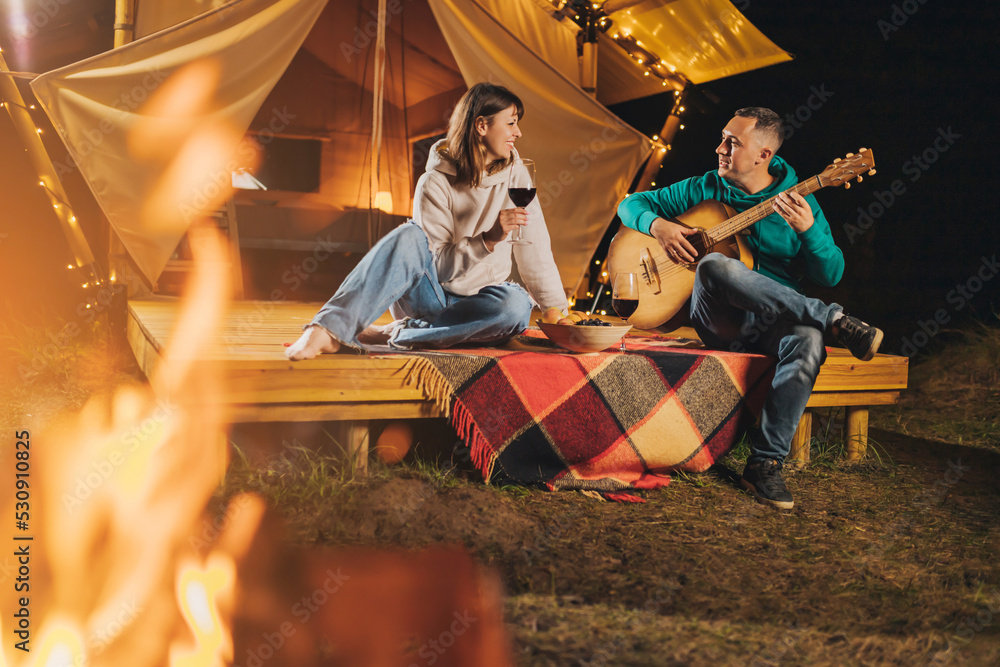 Happy couple relaxing in glamping on autumn evening, drinking wine and playing guitar near cozy bonfire. Luxury camping tent for outdoor recreation and recreation. Lifestyle concept - obrazy, fototapety, plakaty 