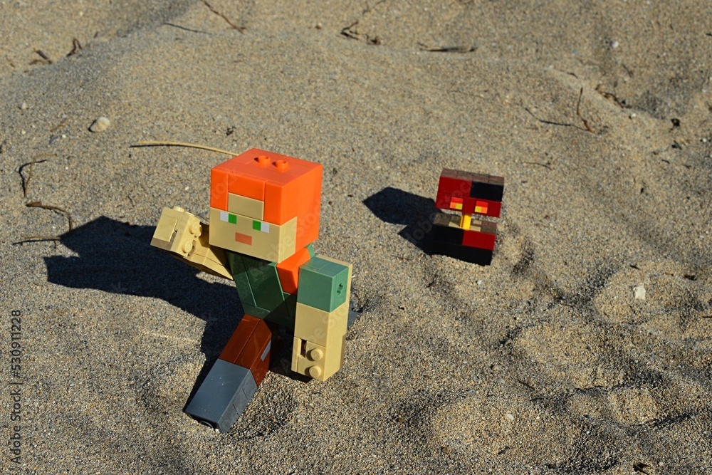 Fototapeta premium LEGO Minecraft large action figure of Alex chased by hostile mob Magma Cube on sandy beach in afternoon sunshine.