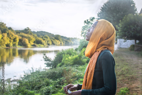 Black woman in hijab looking up at the sky with expression thoughtful outdoors. Peaceful environment in nature. Landscape with river. Copy space. Concept of spirituality. 