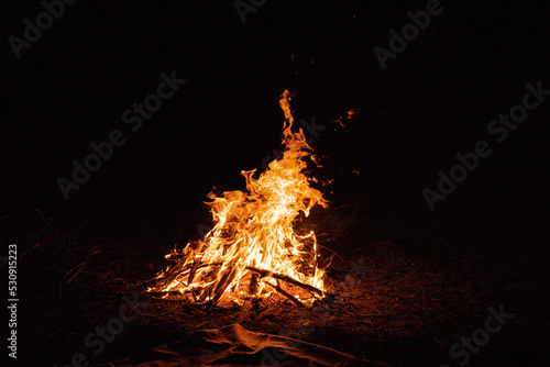 Orange flames fire, remnants bonfire with sparks in forest glade at night near riverside. Charred tree branches. burning out firewood. Big bright flame. Beautiful close-up background, selective focus.