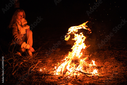 Woman sits near flames fire, remnants bonfire with sparks in forest glade at night near riverside. Charred tree branches. burning out firewood. Big bright flame. Close-up background, selective focus.