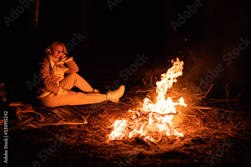 Woman sits near flames fire, remnants bonfire with sparks in forest glade at night near riverside. Charred tree branches. burning out firewood. Big bright flame. Close-up background, selective focus.
