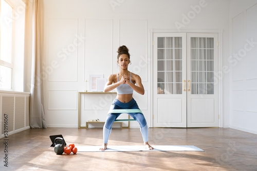 Home Sport. African american woman atching online tutorials on laptop and training on yoga mat in living room. Young girl exercising with fitness elastic bands and enjoying fitness and healthy lifesty