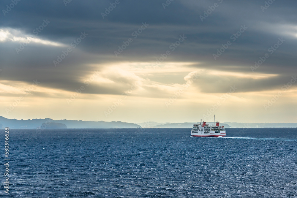 A ferry ship sailing on the sea of the Uraga channel of Tokyo towards the coasts of the Boso peninsula in the morning light below a stormy sky.