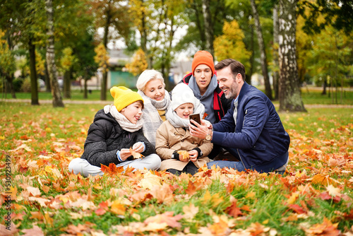 Family of five people spending time and having fun together, taking selfie on the mobile phone or talking online with someone at the autumn golden park.