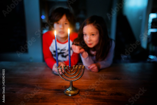 Two siblings sit at table together staring at lit candles on menorah photo