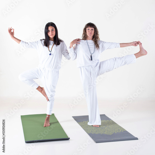 Two women dressed in white doing yoga poses while looking at camera over white background.