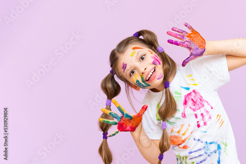 A beautiful little girl stained in multicolored paints on a pink isolated background has fun smiling.