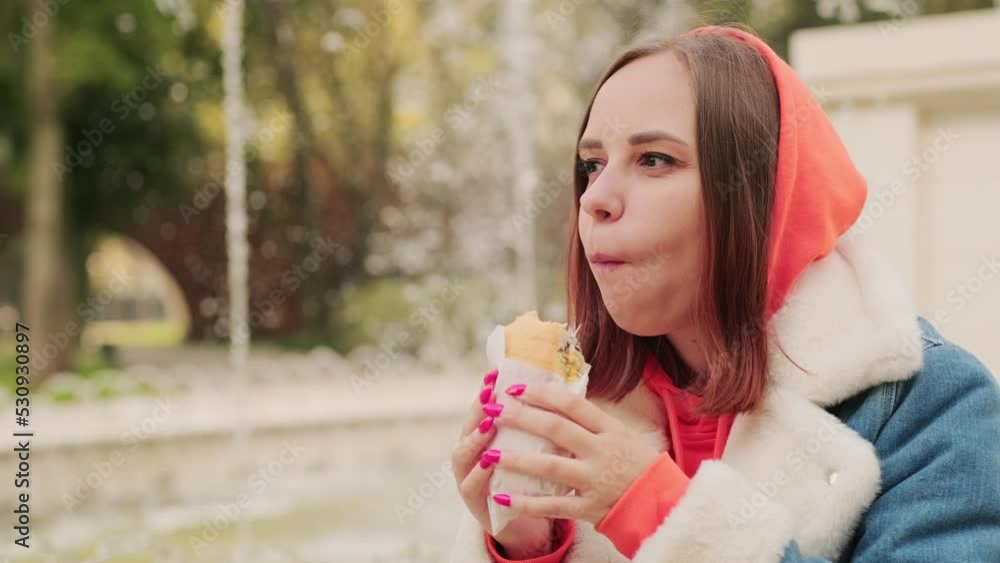 Young positive woman in casual warm clothes eating street food, sitting on fountain in city park. Young positive woman in casual warm clothes eating street food, sitting on fountain in city park.