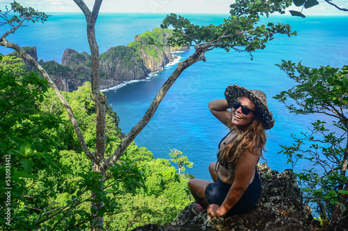 A blonde young woman sitting on Ponta da Sapata Viewpoint and wearing straw hat and sunglasses. Capim açu hiking trail, Fernando de Noronha archipelago, Brazil photo