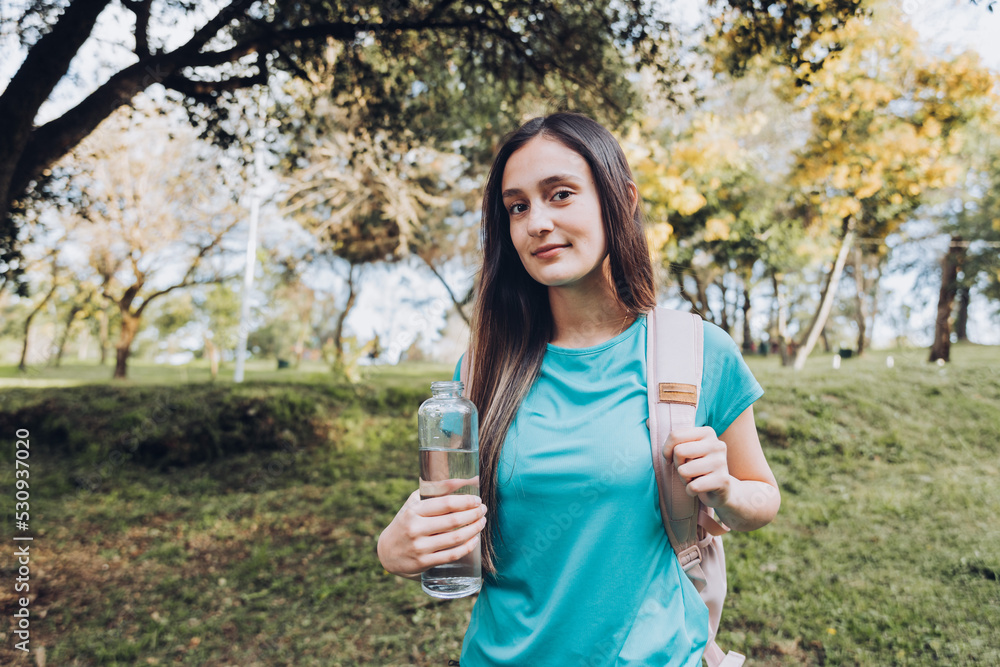 Young girl wearing turquoise t shirt and a pink backpack, holding a glass bottle with water in nature. Natural resource Young girl wearing turquoise t shirt and a pink backpack, holding a glass bottle with water in nature. Natural resource