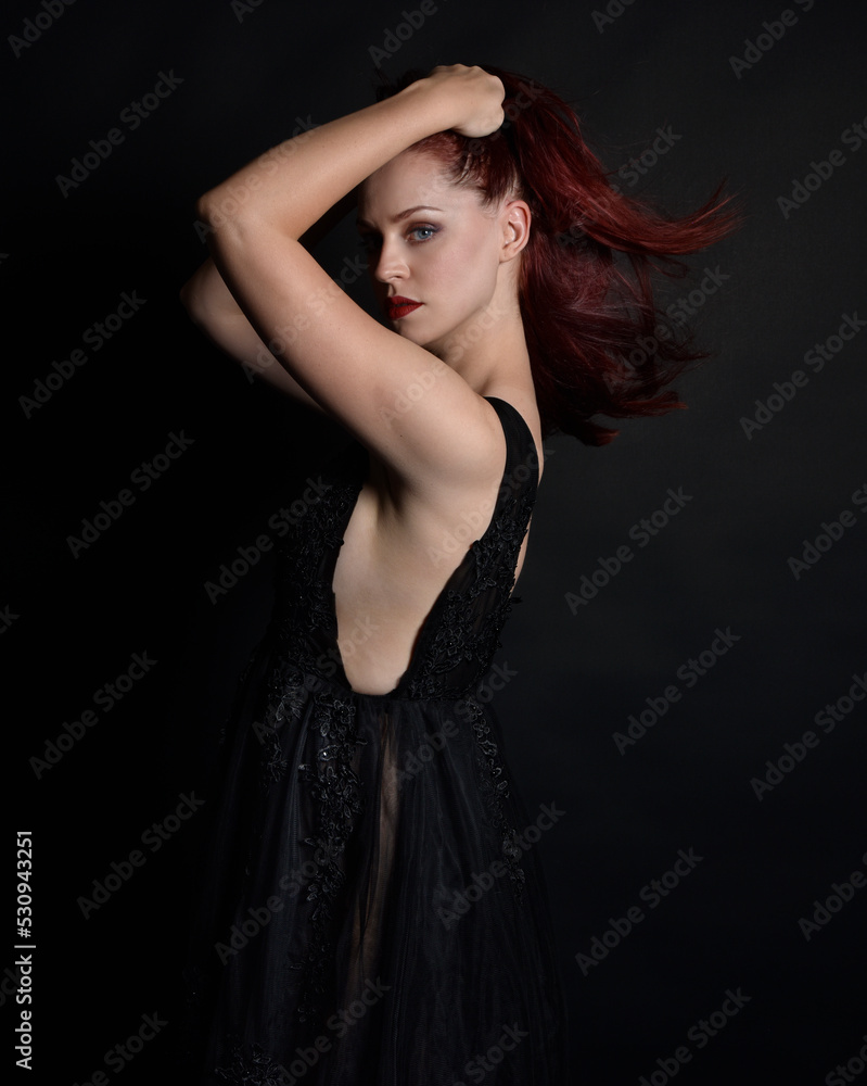 Naklejka premium close up portrait of beautiful woman with red hair, wearing black dress with elegant gestural hand poses on a dark studio background with moody lighting.